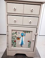 Front view of the small cream painted cupboard with two drawers and painted girl on cabinet door.