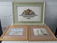 Three framed art pieces. Two beachside prints (children playing on the beach) framed in matching golden frames with mats. One larger shadow box with dried plant arrangement in urn, framed in cream and green mat.