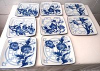 Set of 8 square porcelain plates with blue floral decorations laid out on a surface.