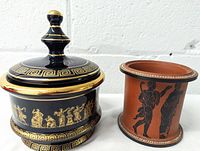 Two vintage Greek lidded dishes side by side, black ceramic with gold and terracotta jar with black figures.