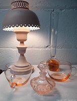 Photo showing the pink oil lamp style electric lamp illuminated with metal shade and glass chimney, amber and clear glass bud vase, pink glass perfume bottle, and pink glass tea cups arranged on table.