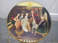 Close-up of the round top tier featuring a decoupaged scene of parlor games with 18th-century dressed figures, detailed gold painted edges around the table top.