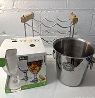 Photo showing Libbey boxed set of 4 wine goblets, metal wine cooler ice bucket in silver metal with handles, on a white surface against a gray wall.