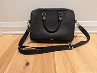 Front view of black leather briefcase showing two handles, shoulder strap, and Boss logo.