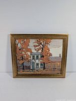 Front view of framed artwork print showing two houses and trees with fall colored leaves in gold toned frame.