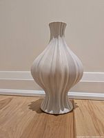 Light grey fluted pottery vase photographed against a beige wall, showing bulbous body and narrow neck with fluted design.