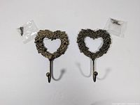 Top down view of two heart-shaped metal hooks with floral detailing and mounting screws in small plastic bags.