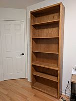 Full view of the tall wooden bookcase with five shelves standing against a white wall near a door.