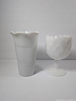 Two vintage white milk glass vases shown side by side on a plain background. The taller vase has a fluted wavy rim and ribbed body. The shorter vase is goblet style with diamond pattern and scalloped rim.