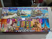 Six Art Gallery 300+ piece puzzle boxes arranged side by side on a table, showing a variety of scenic images and artistic designs.