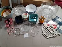Photo of assorted serving items showing pastel nested mixing bowls, pink piggy egg cups, glass bowls, ceramic bowls, and trivets on blue and white checked cloth.