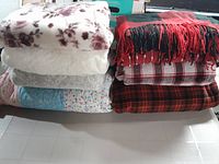 Eight folded blankets and throws stacked in two piles on a tiled floor, showing various patterns and colors including floral, plaid, and solid designs.