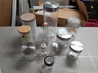 View of assorted canisters and jars including glass jars with metal lids, a white tin canister with wooden lid, and small glass jars with twine strings.