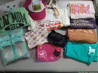 Full lot overview on couch showing shirts stacked, slippers, cap, tote bag, and makeup bags