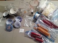 Overview photo showing wax flakes in a bag, multiple glass containers, measuring jug, wax wicks, packaged candles and floral decorative tins.