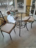 Photo showing the entire round glass top table with two metal framed chairs with beige cushions around it near a window.