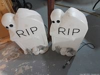 Two white plastic Halloween tombstones side by side with 'RIP' lettering, skulls, and skeletal arm details, cords visible for light-up feature.