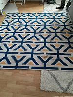 Full view of the flat woven rug laid on a hardwood and carpeted floor, showing its geometric blue, yellow, and white pattern.