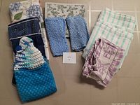 Kitchen linens laid out showing a variety of hand towels, tea towels, and wash cloths including crocheted blue wash cloths, floral and checkered prints.