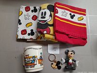 Three dish towels featuring different Mickey Mouse designs, vintage cup with Walt Disney World illustrations, small Mickey figurine and Mickey keychain
