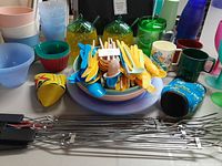 Photo showing colorful plastic bowls, cups, and the yellow plastic cutlery on a purple plate with green pineapple-style drink containers and metal skewers in front.