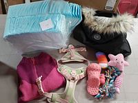Photo showing black parka coat with faux fur hood, pink knit sweater, pink and beige dog harness, pack of blue puppy pads, and multiple colorful chew toys.