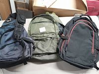 Photo of three backpacks: Quantum brand black/gray nylon backpack with side pockets, olive green backpack, and black with red trim backpack.