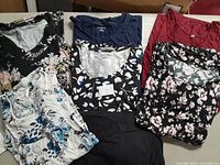 Photo showing six folded plus size shirts and blouses with floral and solid colors including black, navy blue, red, and floral patterns.