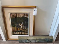 Photo showing two paintings: framed farmhouse painting and long rectangular boat painting leaning on floor
