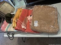 Four blankets stacked: pastel striped, yellow, red/orange, and beige satin trimmed wool blanket with Biltmore hat next to them.