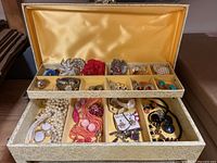 Jewelry box open showing satin-lined top tray with compartments of brooches, earrings and small items