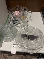 Overview of glass serving items grouped on a table including platters, bowls, glasses in caddy, pitcher and serving utensils.