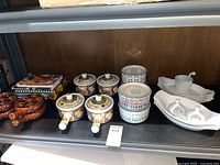 Shelf showing four onion soup bowls with floral lids, three colorful patterned containers with lids, brown ceramic bowls, and cookbooks in the back