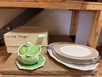 Photo showing a green and white ceramic turkey-shaped platter with leaf design, a small green and white plate underneath, a stack of white and gold rim plates, and a box labeled '10 Gold Chargers'.