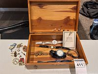 Wooden jewelry box containing 5 wrist watches, assorted cuff links, and brooches on a white surface.