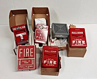 Top view of five red fire alarm pull stations, three in boxes and two used, plus gray Federal fire horn new in box.