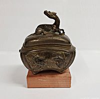 Front view of antique metal container with dragon lid and dragon side decorations, mounted on a wooden block for support, showing wear and patina.