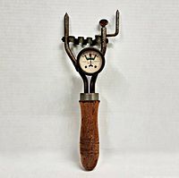 Full view of battery tester showing wooden handle, metal body, prongs, and analog gauge.