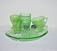Full lot of 8 pieces showing the green glassware including cups, salt and pepper shakers, tumbler, footed glass, and oval dish glowing under white light