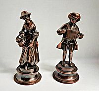 Front view of the two copper-colored metal figurines, a lady holding a basket and a man with an accordion, on round bases.