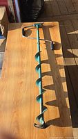 Full view of the green manual ice auger on a wooden surface, showing the handle and spiral drill bit.
