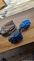 Three baseball gloves on a wooden table: a brown Wilson adult glove and two kids' gloves, one blue Wilson and one black Rawlings.