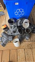 Blue recycling bin filled with various ABS plumbing fittings showing multiple elbow, tee, and coupling pieces in black and some white fittings, primarily 3" and 4" sizes.
