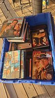 Top view of a blue bin containing various DVD movies including 'License to Wed,' 'Pirates of the Caribbean,' and 'Indiana Jones and the Last Crusade.'