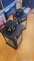Two black metal hanging candle lanterns on wooden surface; each has a square base and glass sides, with a handle on top.