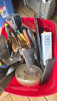 Mixed cement, tile, drywall tools in red plastic bin, showing various metal trowels, putty knives, scrapers with different colored handles and a large roll of tape