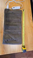 Full heating pad laid flat next to tape measure showing approximately 20 inches length, brown color with printed instructions on fabric surface.