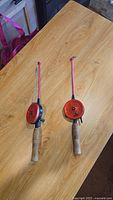Photo showing two ice fishing rods with red reels and cork handles laid on wooden table.
