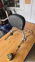 Photo of black padded motorcycle backrest with chrome luggage rack and mounting hardware on wooden table, tape measure visible showing dimensions of mounting bracket.