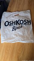 White plastic shopping bag with OshKosh B'Gosh logo printed in dark blue, cutout handles visible, laid flat on wood surface